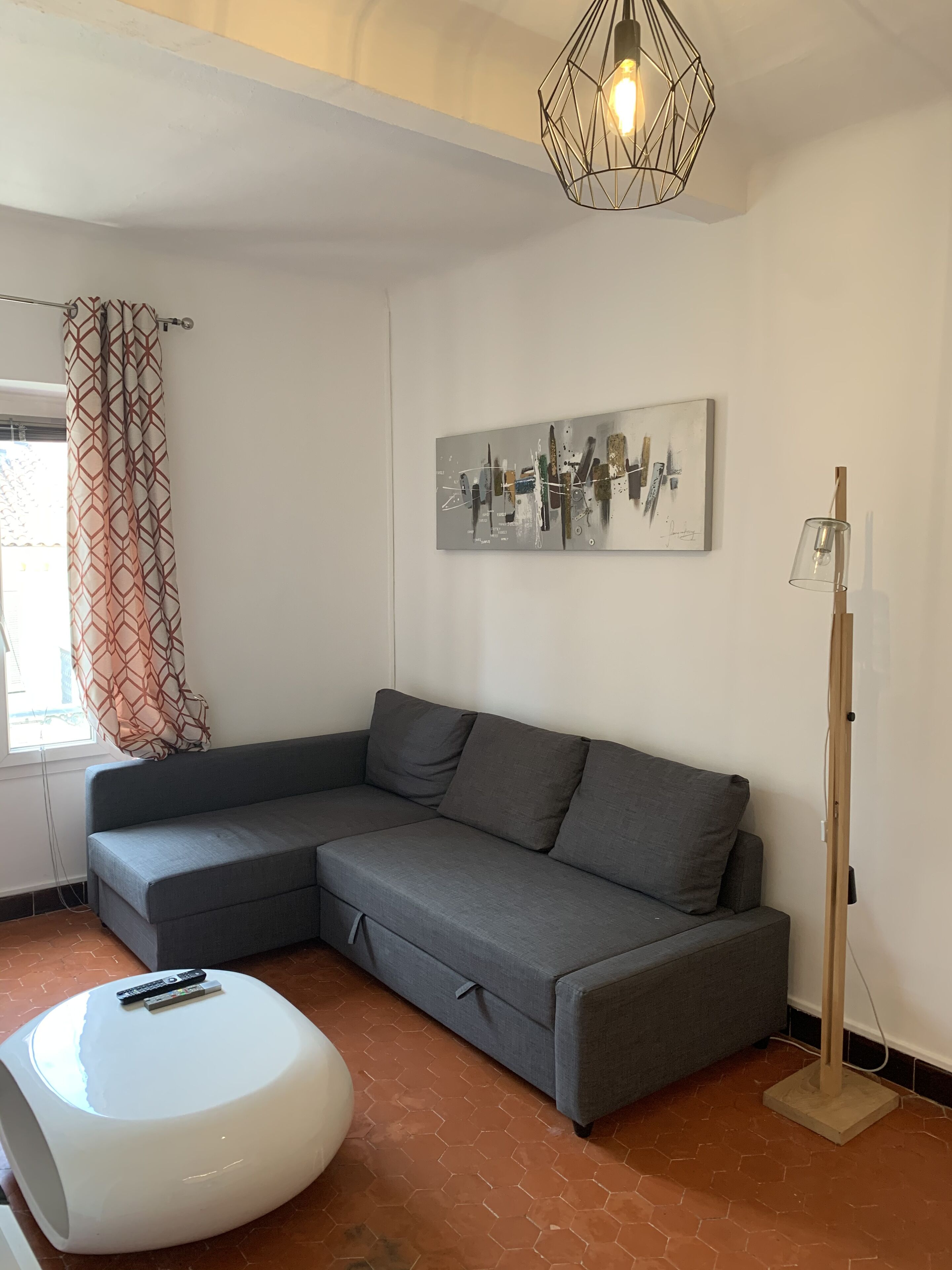 A modern living room with a grey sectional sofa, a white ottoman, a geometric pendant light, a floor lamp, and a piece of abstract art on the wall.