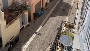 A narrow cobblestone street lined with pastel-colored buildings, a scooter parked on the sidewalk, and a small table with a chair on the balcony.