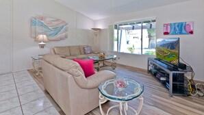 Una sala de estar con un sofá beige, una mesa de centro de vidrio, una lámpara y un mueble para televisión con libros.