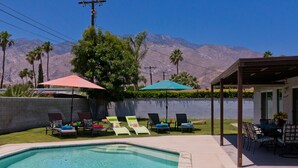 Una piscina con sillas de descanso y sombrillas, un patio cubierto y montañas al fondo.