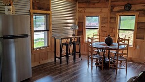 Interior de una cabaña de madera con una zona de cocina, que incluye un refrigerador, una mesa de comedor con sillas y una ventana con persianas.