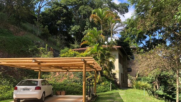 A white car parked under a wooden pergola in a lush garden with a house in the background. - Retreat with Suite in a Secure Condominium | Nature and Stunning Views  (RJ)
