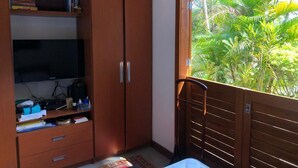 A bedroom with a wooden wardrobe, a flat-screen TV, and a window with a view of tropical foliage. - Retreat with Suite in a Secure Condominium | Nature and Stunning Views  (RJ)