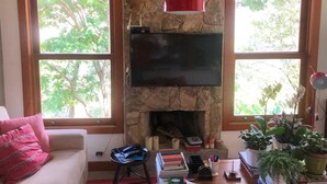 A living room with a stone fireplace, a flat-screen TV, and large windows with a view of trees. - Retreat with Suite in a Secure Condominium | Nature and Stunning Views  (RJ)