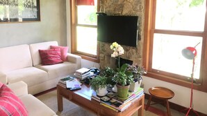 A living room with a stone wall, a flat-screen TV mounted above a fireplace, a sofa with a pink pillow, a coffee table with books and plants, and a red pendant light. - Retreat with Suite in a Secure Condominium | Nature and Stunning Views  (RJ)