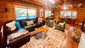 A cozy log cabin interior with a leather sofa, green armchairs, a patterned rug, and a stone fireplace. - The Creek-Side Cabin: Hot Tub, Fire Pit,5 Star, AC, Big Deck, Quiet,Walk To Town (Idyllwild-Pine Cove)