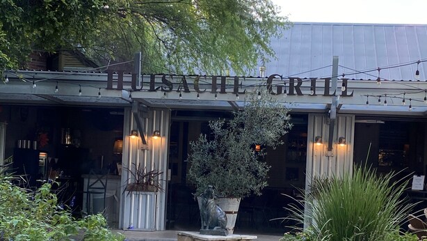Um restaurante chamado "Isabelle Grill" com um caminho de cascalho que leva à entrada, rodeado por vegetação e plantas em vasos.