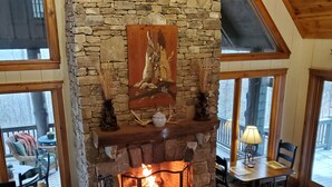 A cozy living room with a stone fireplace, comfortable seating, and a view through large windows.