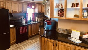 A kitchen with wooden cabinets, a black refrigerator, and a black countertop. There is a window with red and black curtains, and a sign that says "Bless Our Home".