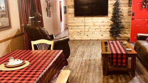 A cozy cabin interior with a dining area, a sofa, a small table, and a television mounted on a wooden wall.