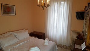 Quarto de hotel com cama, mesa de cabeceira, lustre, janela com cortinas, radiador e televisão na parede.