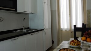 Cozinha com armários brancos, bancada preta, pia de aço inoxidável e micro-ondas. Há uma janela com cortinas leves e uma mesa com uma fruteira e uma torta.