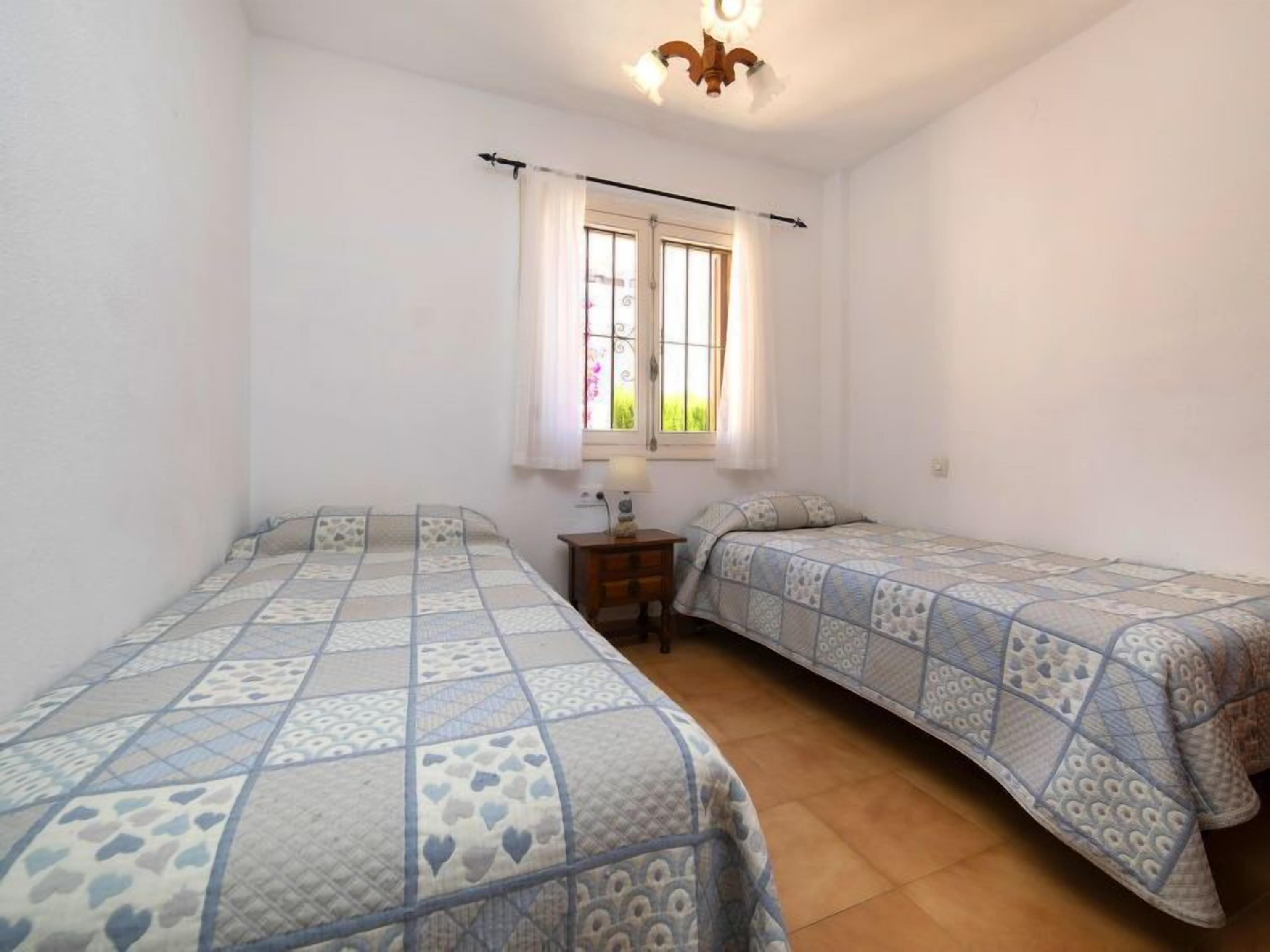 A small, clean room with two single beds, a window with curtains, and a small table with a lamp.
