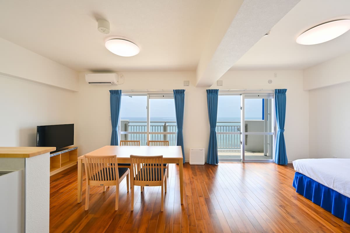 A hotel room with a wooden floor, a bed with a blue skirt, a dining table with chairs, a television, and a balcony with blue curtains.