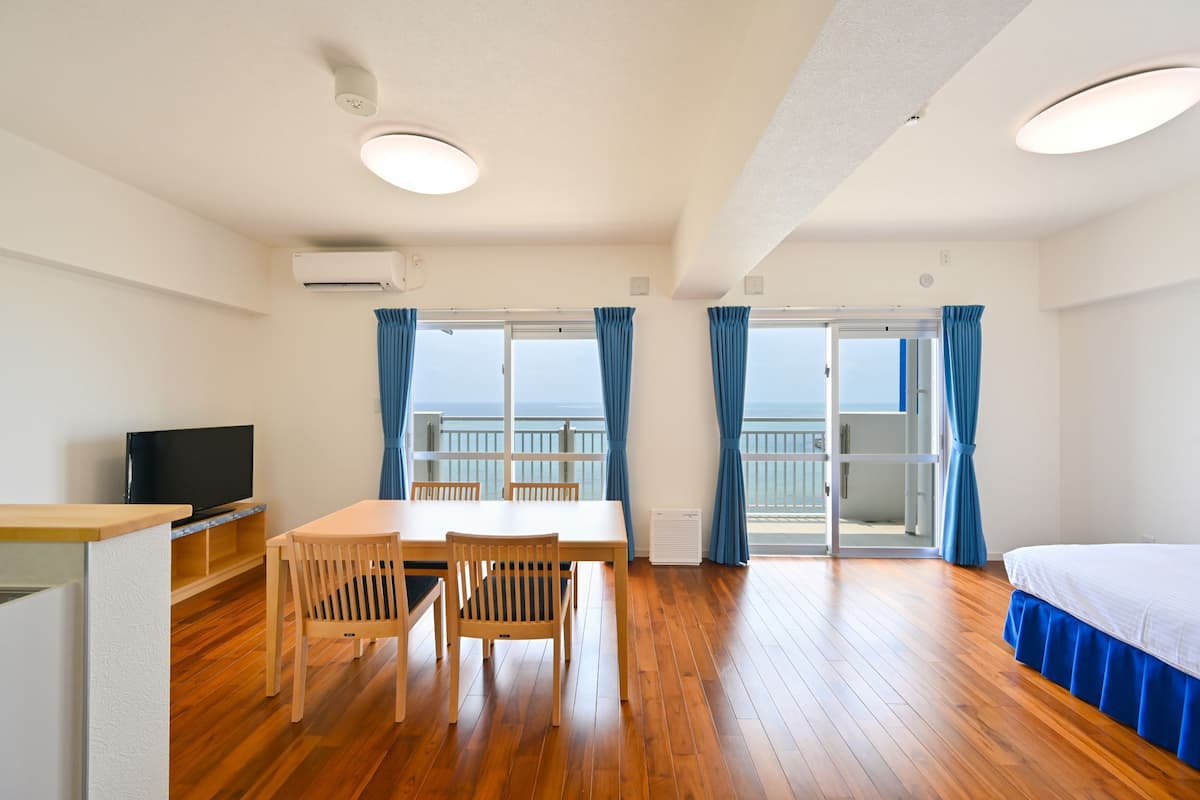 A hotel room with a wooden floor, a dining table with chairs, a TV, a bed, and a balcony with curtains.