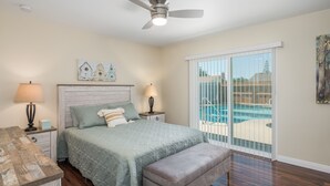 Un dormitorio con cama, mesita de noche, banco, ventilador de techo y vista a una piscina a través de una puerta corrediza de vidrio.