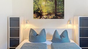 Una camera da letto con due letti, un quadro appeso raffigurante una foresta, lampade sui comodini e mobili contenitori a muro.