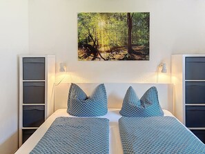 Ein Schlafzimmer mit zwei Betten, einem an der Wand befestigten Bild von einem Wald, Nachttischlampen und eingebauten Stauraumelementen.