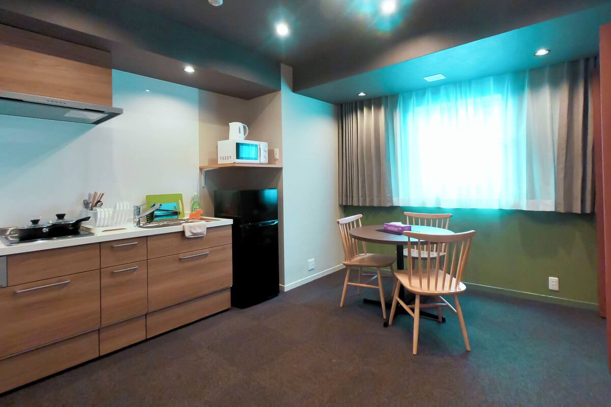 A compact kitchen with a microwave, stove, and refrigerator. There is a dining table with chairs and a window with curtains.