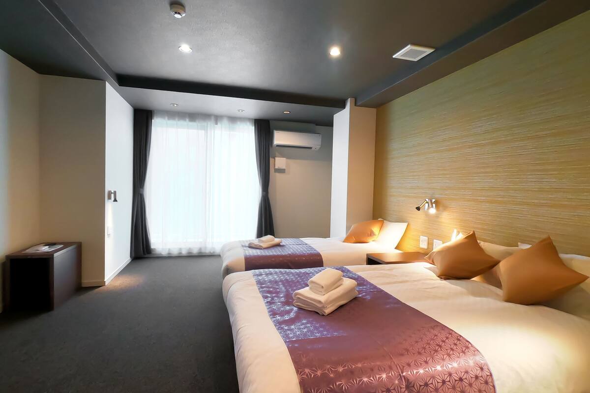 A modern hotel room with two beds, a large window with curtains, an air conditioning unit, and a wall-mounted light fixture.