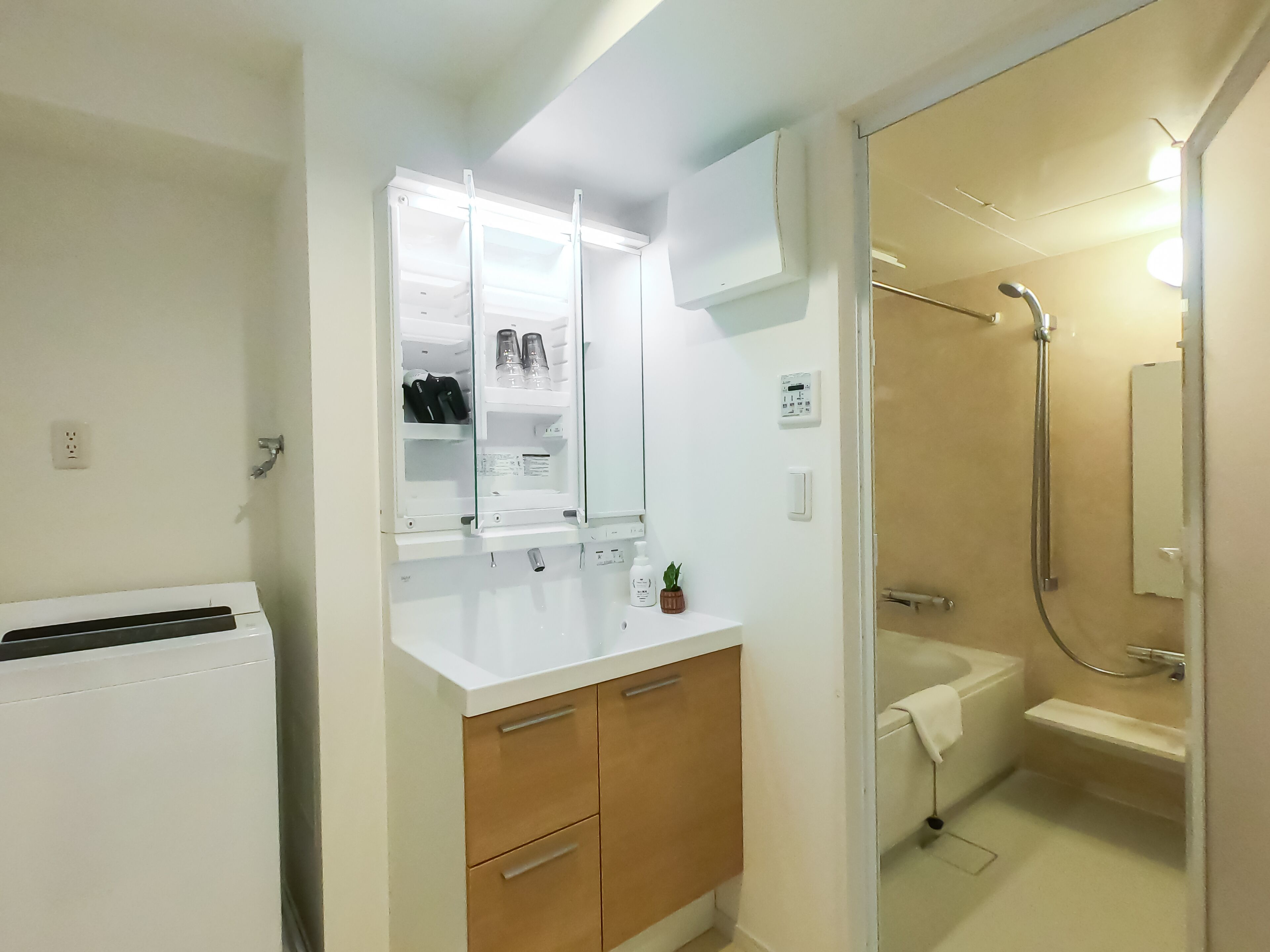 Un baño pequeño con un lavamanos blanco, armarios de madera, una cabina de ducha de vidrio y un enchufe a la vista.