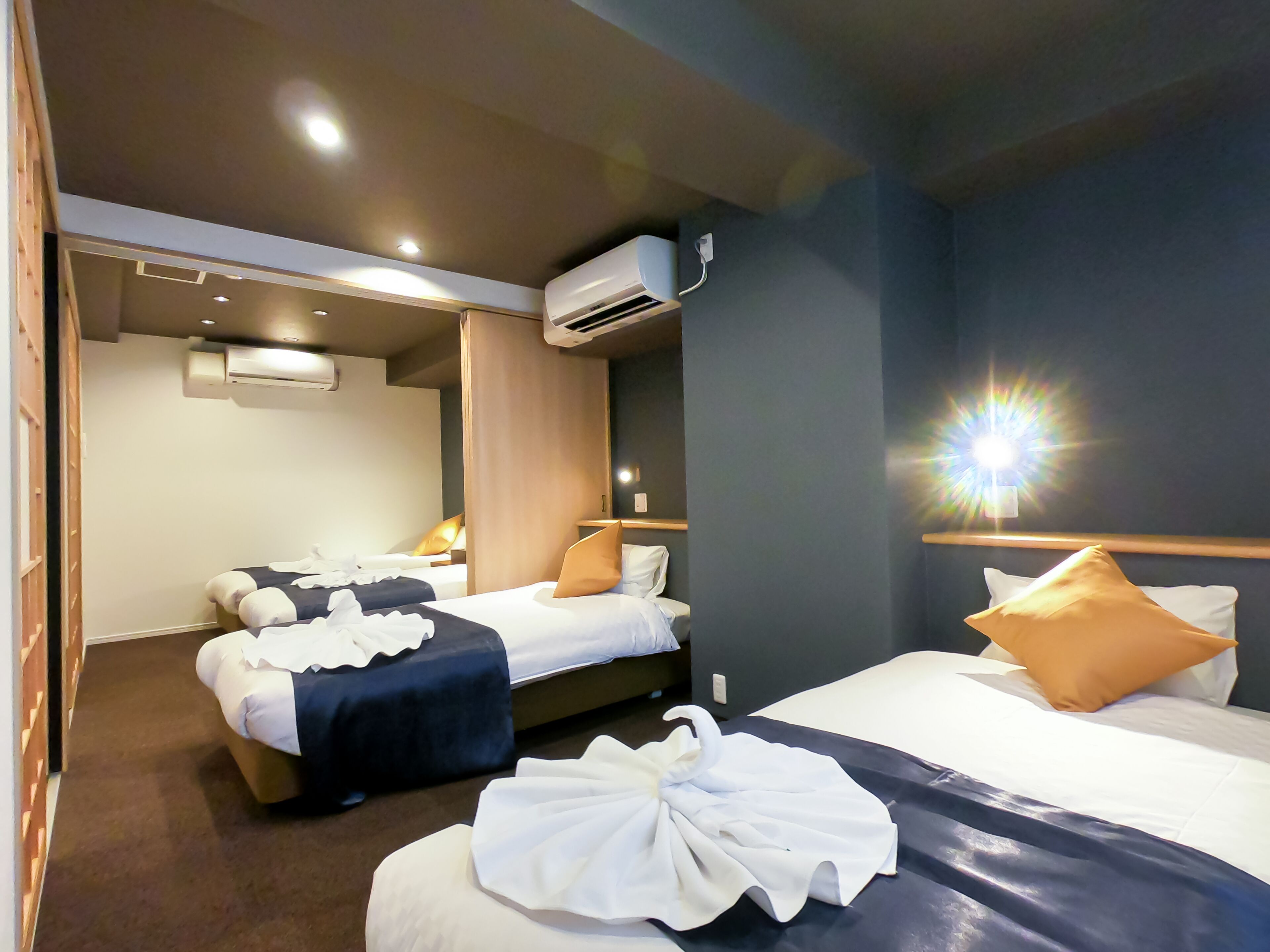 A hotel room with two beds, a ceiling fan, an air conditioning unit, and a wall-mounted light fixture.