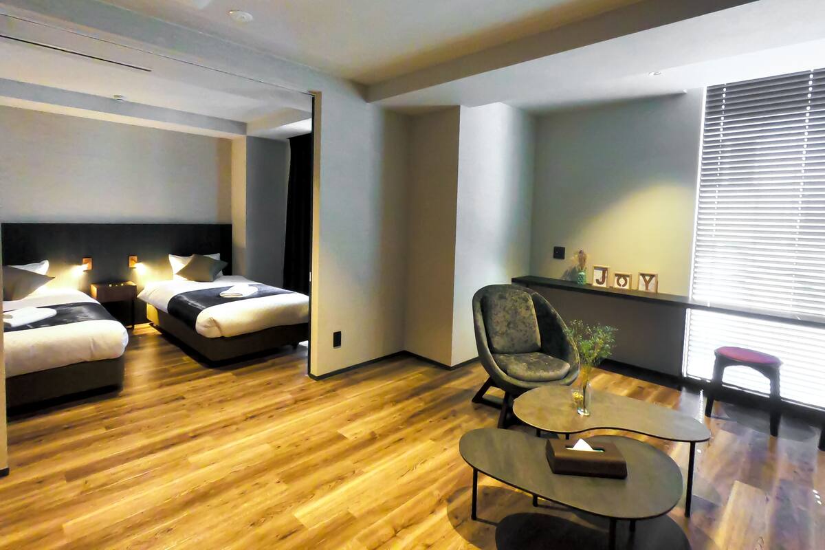 A modern hotel room with two beds, a small table, a chair, and a large window with blinds.