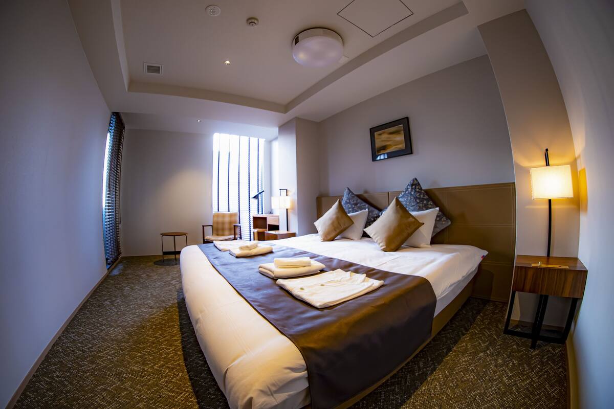 A hotel room with a large bed, a bedside table with a lamp, a window with blinds, and a carpeted floor.