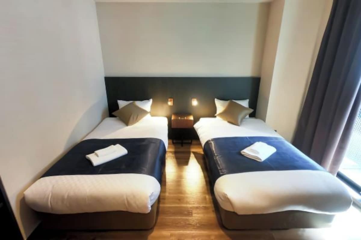 Habitación de hotel con dos camas individuales, cada una con una almohada y una toalla doblada.