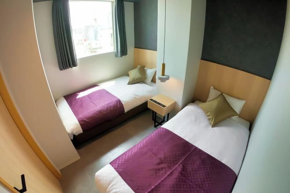 Hotellihuone, jossa on kaksi sänkyä, yöpöytä, ikkuna verhoilla ja näkymä kaupunkiin.