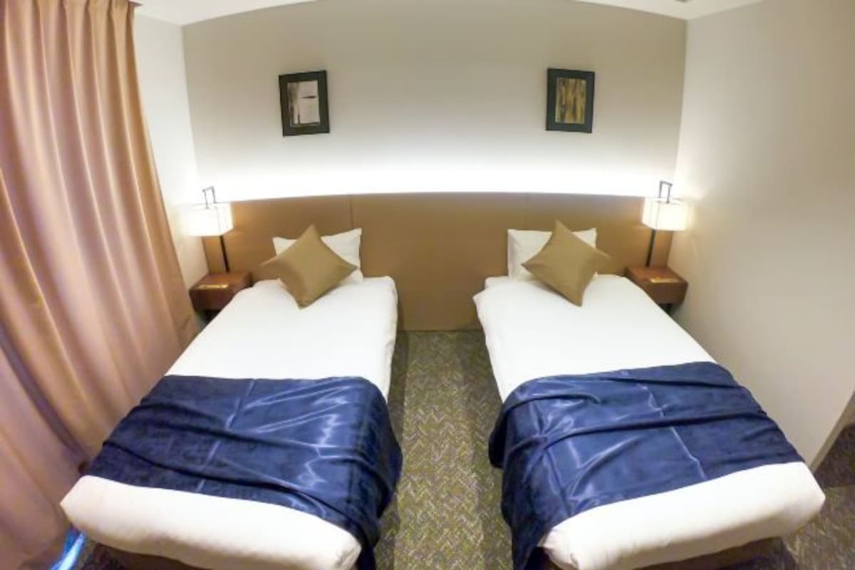 Habitación de hotel con dos camas, cada una con cabecera, mesitas de noche, lámparas y una cortina a un lado.