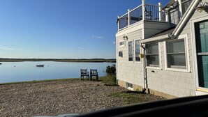 A house with a balcony overlooking a calm lake, two chairs on the gravel, and a clear sky.