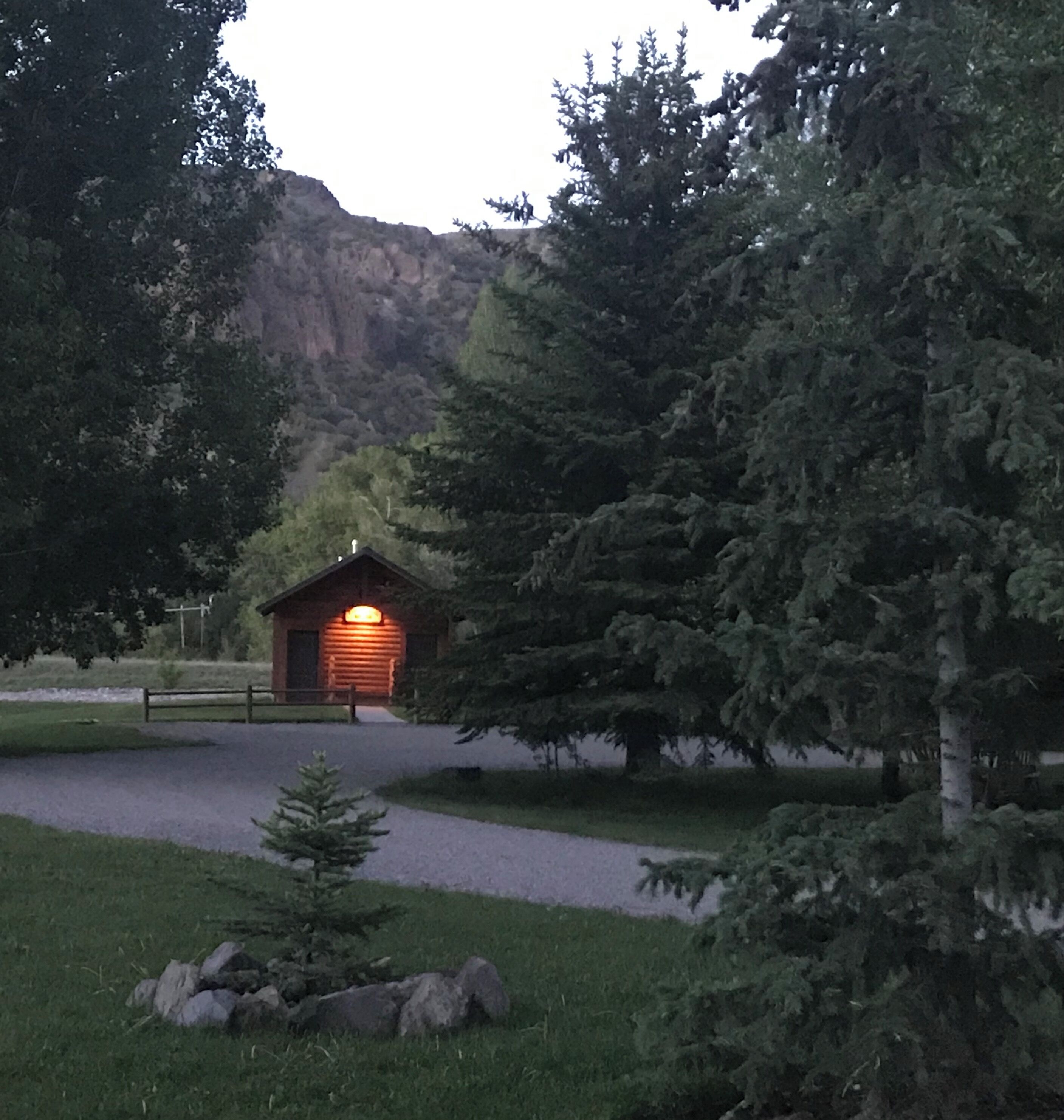 Uma pousada com fachada de madeira e entrada iluminada, rodeada por uma floresta densa e um cenário montanhoso.
