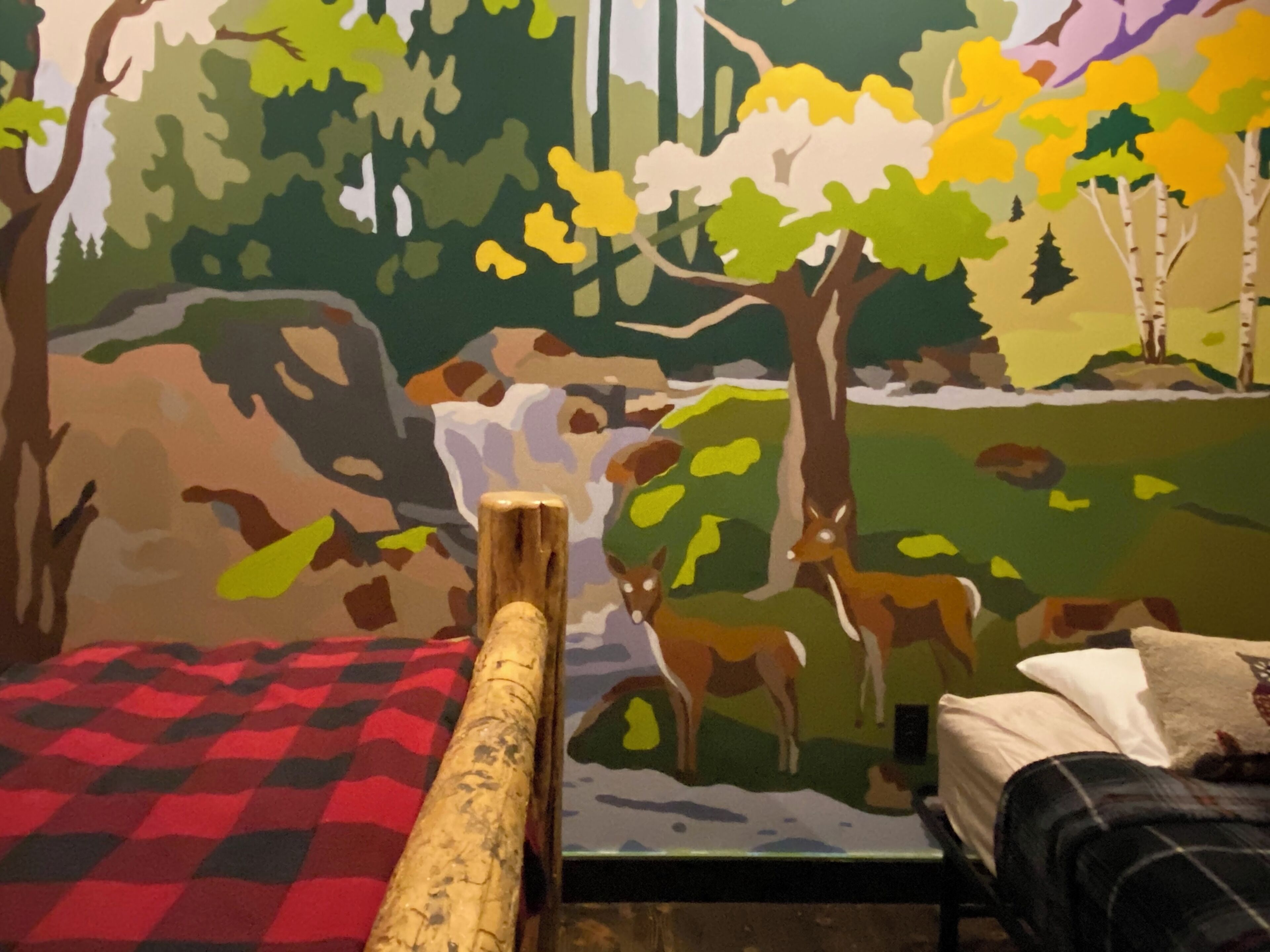 Una habitación con una cama, una colcha de cuadros rojos y negros y un mural con una escena de bosque.