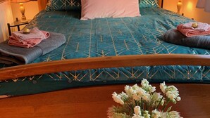 Een net opgemaakt bed met een blauwgroene spreidek en decoratieve kussens, een bloevas op een tafel en twee lampen aan weerszijden van het bed.