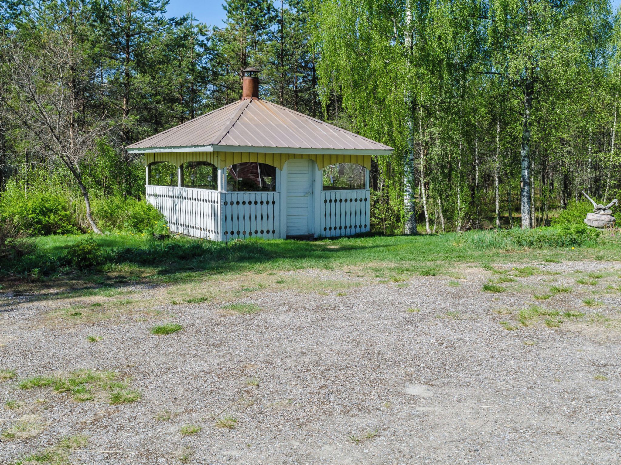 Un petit kiosque doté d’un toit en métal et de murs blancs, entouré d’arbres et bordé d’un chemin de gravier.