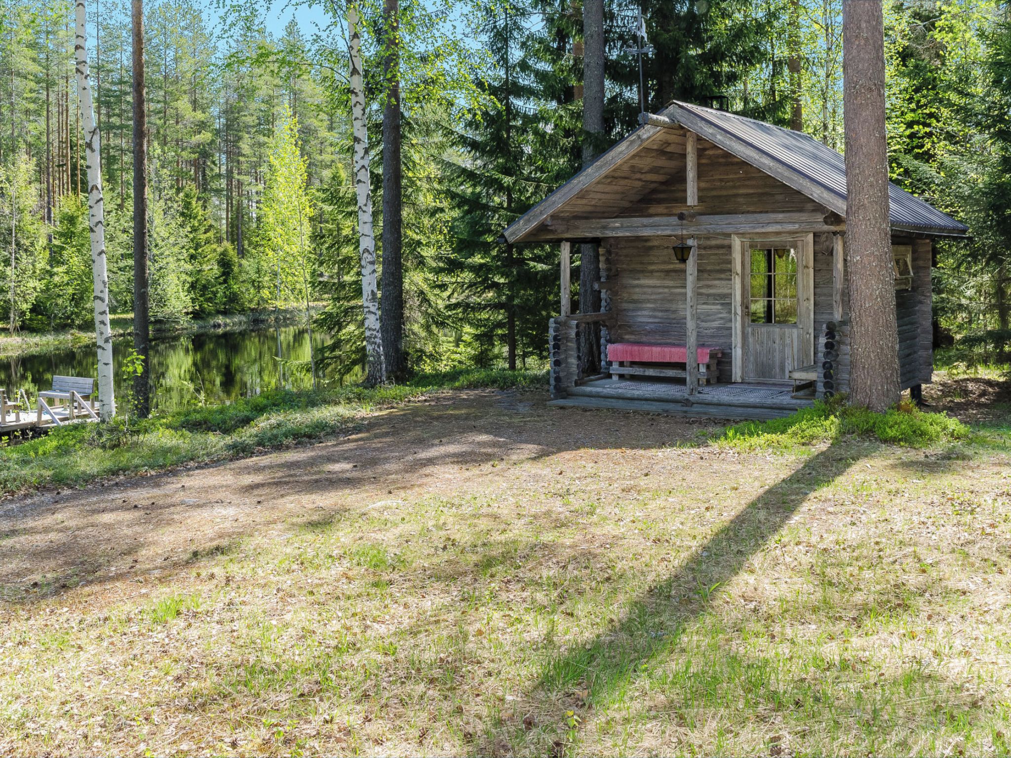 A rustic cabin with a porch and a bench, surrounded by a forest and a lake.