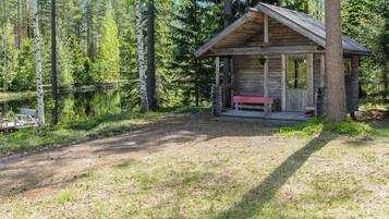 A rustic cabin with a porch and a bench, surrounded by a forest and a lake.