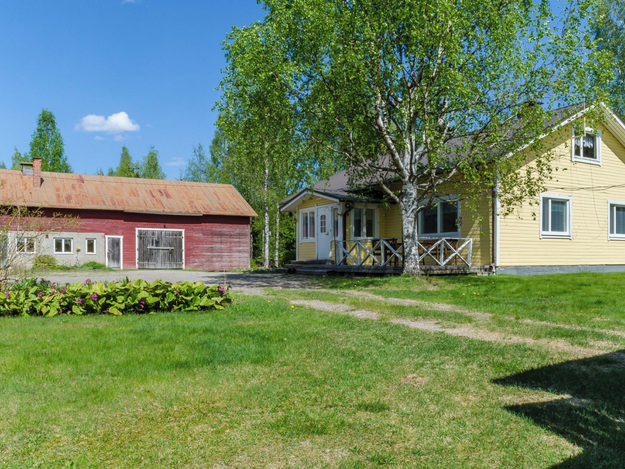 Maaseudulla sijaitseva kiinteistö, jossa on punainen lato, keltainen talo kuistilla ja suuri puu etualalla.