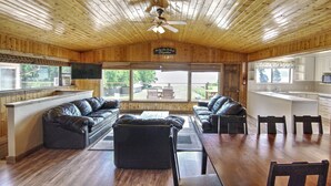 A spacious living area with a wooden ceiling, a large window, a fireplace, and a kitchen area with white cabinets and a countertop.