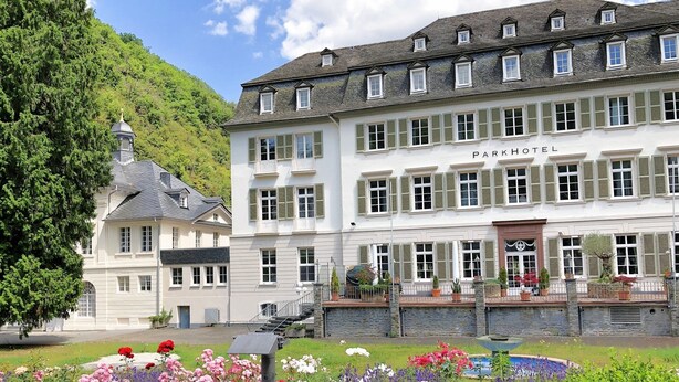 Um hotel grande, de vários andares, com arquitetura europeia clássica, apresentando uma entrada imponente com escadaria e uma pequena torre.