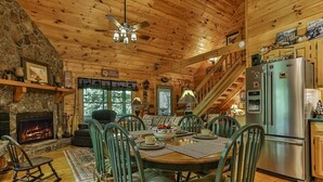 A wooden cabin interior with a stone fireplace, a dining table with chairs, a kitchen area, and a staircase.