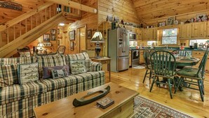 A wooden cabin interior with a sofa, chairs, a dining table, a kitchen area, and a staircase.