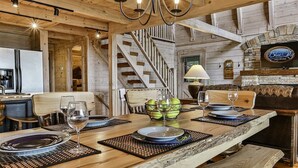 A rustic wooden interior with a dining table set for two, a staircase, and a television mounted on the wall.