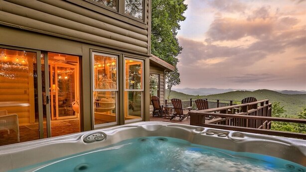 A hot tub on a deck with a view of hills, a covered patio with chairs, and a building with glass doors.