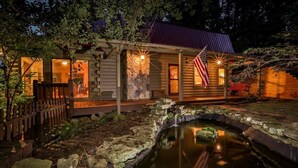 A rustic cabin with a stone foundation, a small pond, and an American flag.