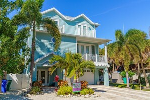 A two-story house with a balcony, surrounded by tropical plants and a clear blue sky. - Big Family Beach House: Large family home, heated pool, putting green (Anna Maria)