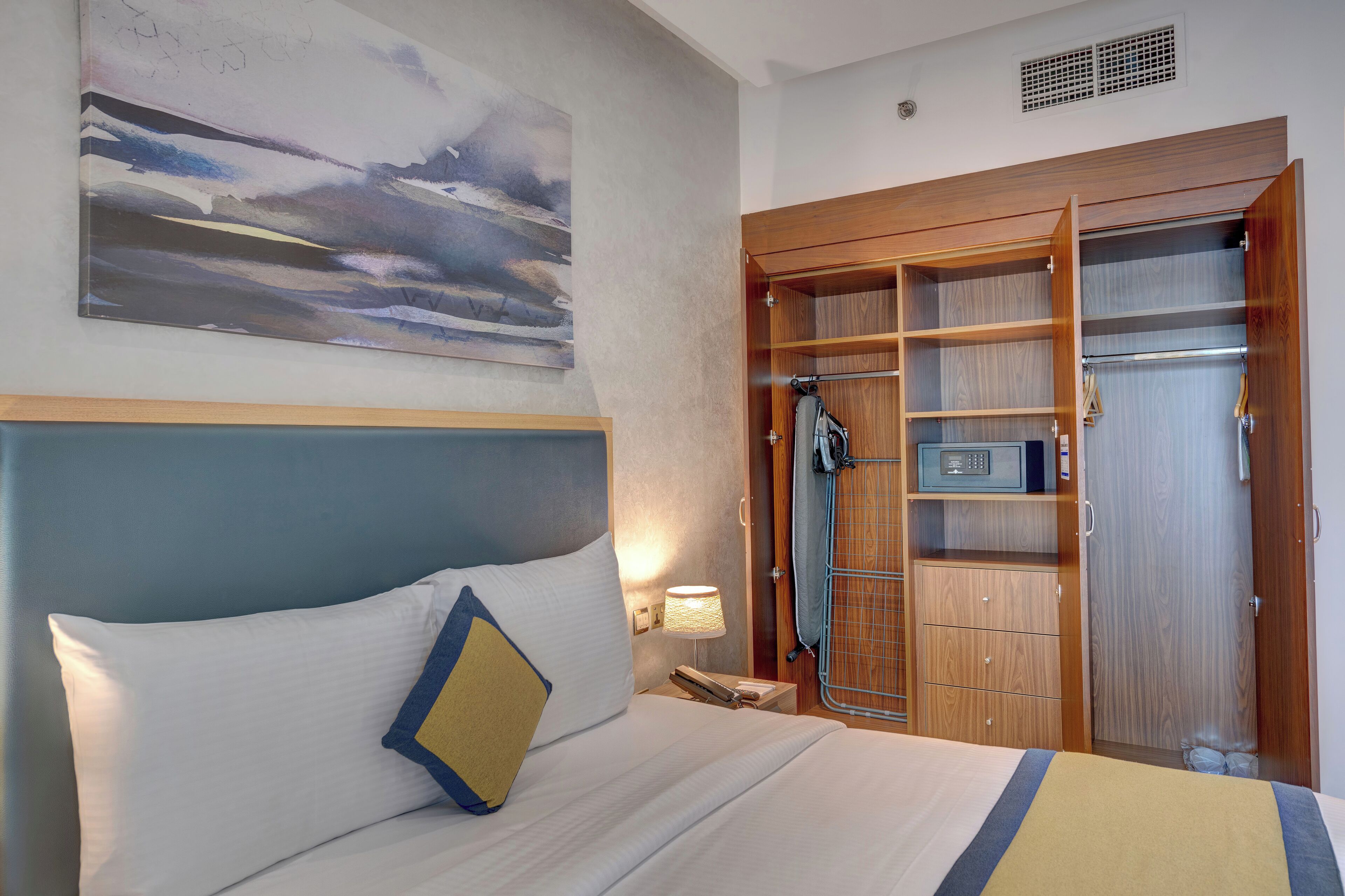 Une chambre d’hôtel comprenant un lit, une table de chevet, une lampe, un tableau, une armoire en bois et un espace douche.