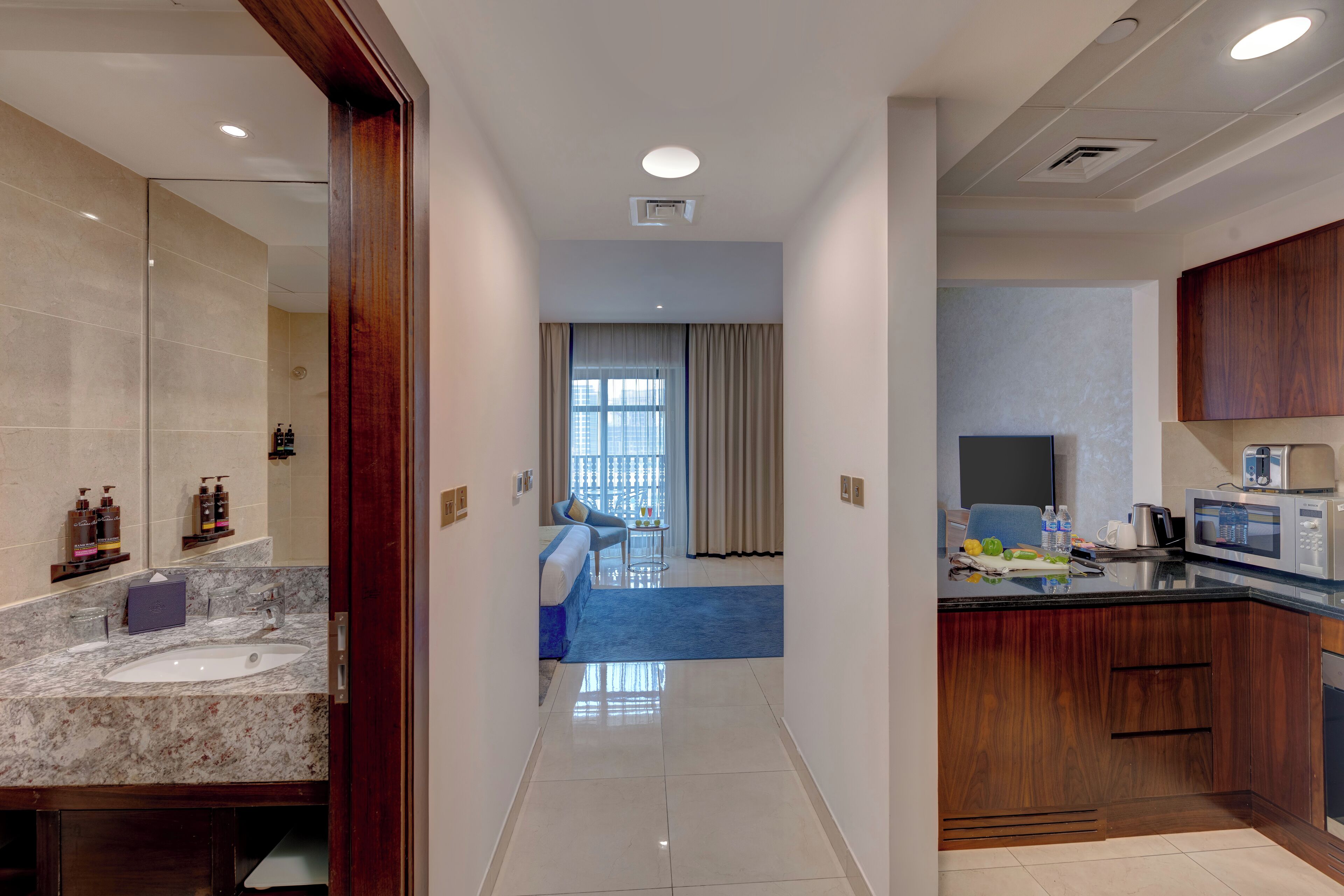 A modern hotel room with a marble bathroom, a kitchenette, a dining area, and a view of the city through large windows.
