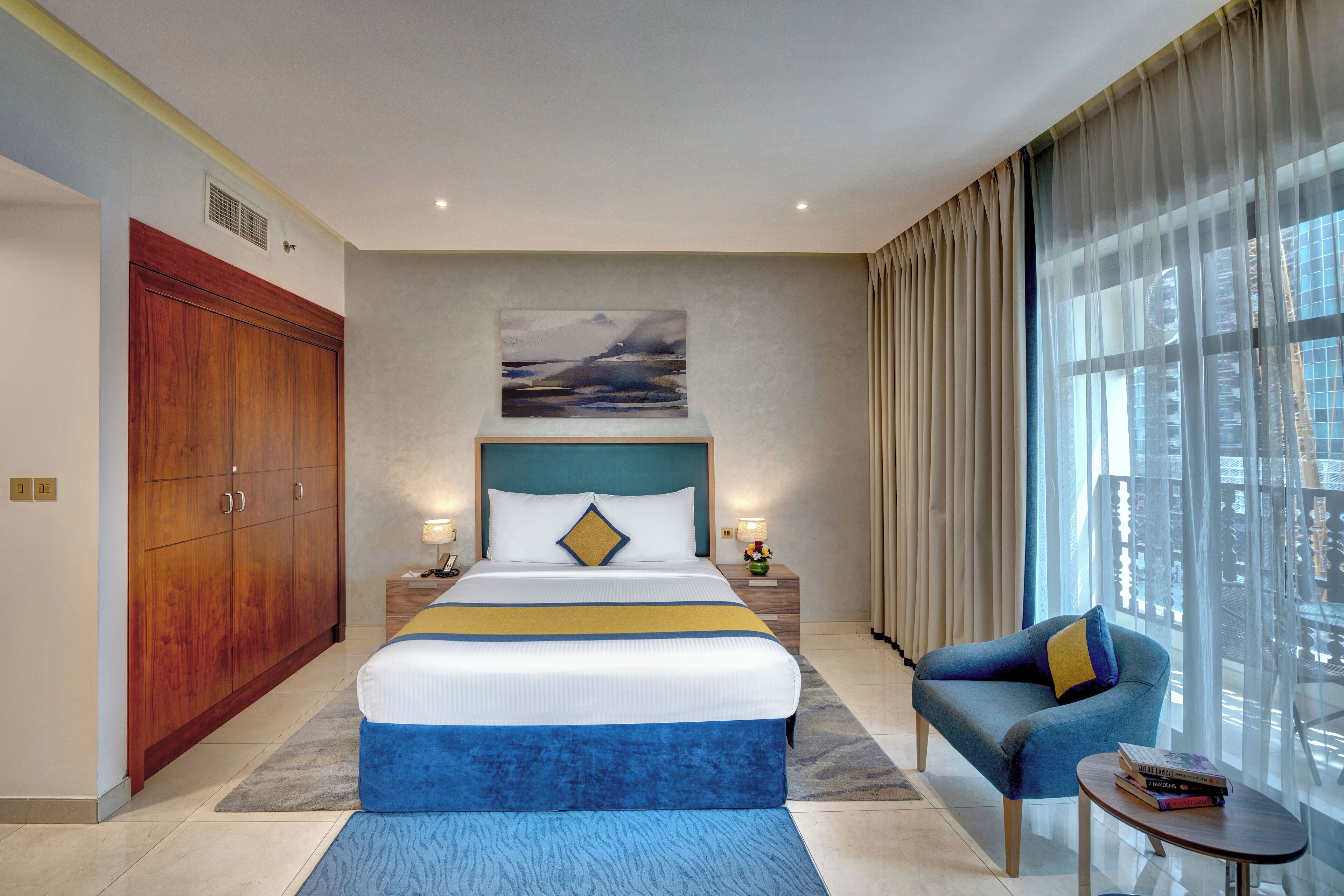 A hotel room with a large bed, a wooden wardrobe, a blue sofa, a small table with books, and a large window with curtains.